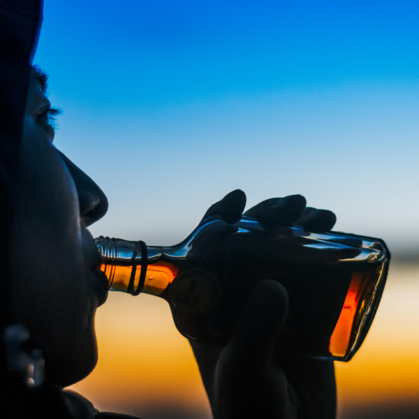 Eine Person in einer Kapuzenjacke trinkt bei Sonnenuntergang aus einer Glasflasche, während der Himmel im Hintergrund von tiefem Blau zu Orange übergeht.