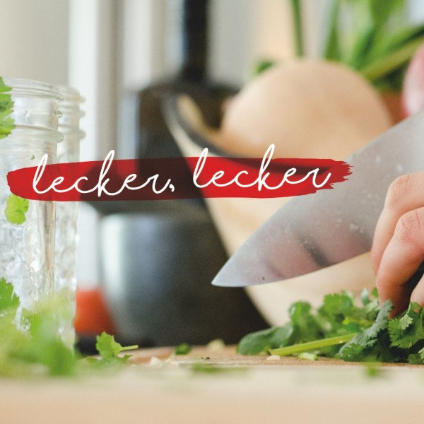Eine Hand mit einem Ring hackt frischen Koriander auf einem Schneidebrett mit einem Messer. Im Hintergrund befinden sich Gemüse und ein Glasgefäß. Der Schriftzug lecker, lecker erscheint in Weiß auf einem roten Pinselstrich.