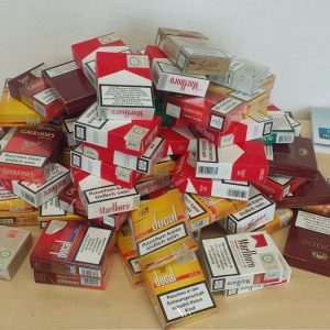 Ein großer Stapel leerer Zigarettenschachteln, hauptsächlich Marlboro und Gauloises, liegt auf einem Holztisch ausgebreitet. Um den Stapel herum liegen Prospekte und Broschüren über die Raucherentwöhnung.