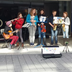 Sechs junge Musiker treten auf einem Bürgersteig auf; sie spielen Gitarre, Flöte, Geige, Saxophon und Klarinette. Vor ihnen steht ein Schild und ein offener Kasten für Spenden. Die Gruppe steht in der Nähe eines großen Gebäudes mit Fenstern.