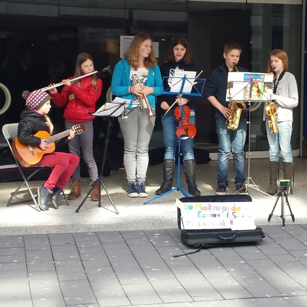 Sechs junge Musiker treten auf einem Bürgersteig auf; sie spielen Gitarre, Flöte, Geige, Saxophon und Klarinette. Vor ihnen steht ein Schild und ein offener Kasten für Spenden. Die Gruppe steht in der Nähe eines großen Gebäudes mit Fenstern.