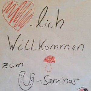 Ein Plakat mit dem deutschen Text Herzlich Willkommen zum U-Seminar, verziert mit einem roten Herz, einem Pilz, einem Hufeisen, einem vierblättrigen Kleeblatt und einem Marienkäfer, alle mit bunten Markern gezeichnet.