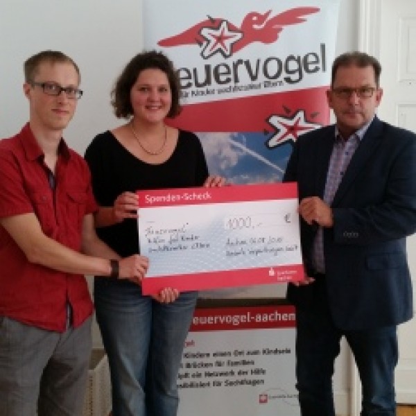 Drei Personen stehen in einem Haus und halten einen großen Spendenscheck über 1.000 Euro vor einem Schild mit der Aufschrift Feuervogel. Sie lächeln und schauen in die Kamera.