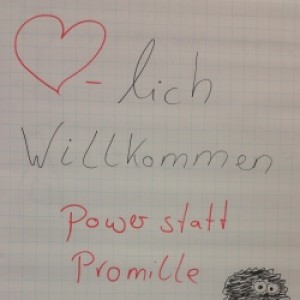 Ein Flipchart mit handgeschriebenem Text: Herzlich Willkommen. Power statt Promille. Yvonne Michel Suchthilfe Aachen. Ein rotes Herzsymbol und eine kleine Zeichnung einer schwarz-weißen Eule in Cartoonform.