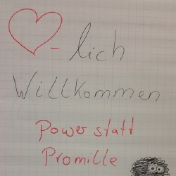 Ein Flipchart mit handgeschriebenem Text: Herzlich Willkommen. Power statt Promille. Yvonne Michel Suchthilfe Aachen. Ein rotes Herzsymbol und eine kleine Zeichnung einer schwarz-weißen Eule in Cartoonform.