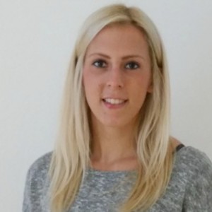 Eine Frau mit glattem, langem blondem Haar und einem grauen Pullover steht vor einem schlichten weißen Hintergrund und lächelt leicht in die Kamera.