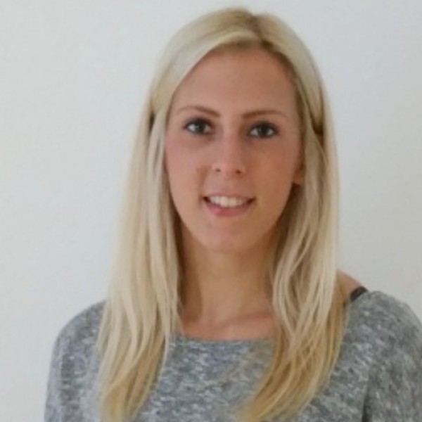 Eine Frau mit glattem, langem blondem Haar und einem grauen Pullover steht vor einem schlichten weißen Hintergrund und lächelt leicht in die Kamera.