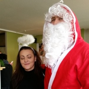 Eine als Weihnachtsmann verkleidete Person steht neben einer Frau, die ein Stirnband mit Heiligenschein trägt, was auf ein Engelskostüm schließen lässt. Die Frau hält ein Klemmbrett in der Hand und lächelt mit geschlossenen Augen. Sie scheinen sich in einem Haus zu befinden.