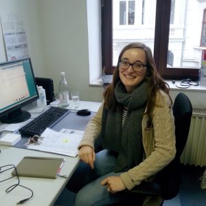 Eine Frau mit langen braunen Haaren, einer Brille und einem grauen Schal sitzt lächelnd an einem Schreibtisch mit einem Computer, Papieren und Büromaterial in einem hellen Raum mit einem Fenster, das Gebäude nach draußen zeigt.
