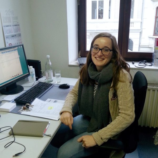 Eine Frau mit langen braunen Haaren, einer Brille und einem grauen Schal sitzt lächelnd an einem Schreibtisch mit einem Computer, Papieren und Büromaterial in einem hellen Raum mit einem Fenster, das Gebäude nach draußen zeigt.