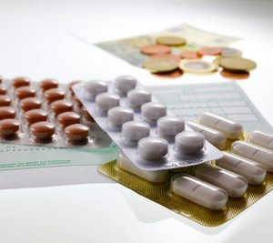 Blisterpackungen mit verschiedenen Pillen und Tabletten sind auf einem Tisch angeordnet, wobei Rezeptformulare und Euro-Münzen im Hintergrund verschwimmen, was auf das Thema Medikamentenkosten oder Ausgaben im Gesundheitswesen hindeutet.
