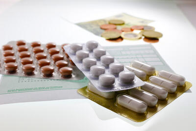 Blisterpackungen mit verschiedenen Pillen und Tabletten sind auf einem Tisch angeordnet, wobei Rezeptformulare und Euro-Münzen im Hintergrund verschwimmen, was auf das Thema Medikamentenkosten oder Ausgaben im Gesundheitswesen hindeutet.