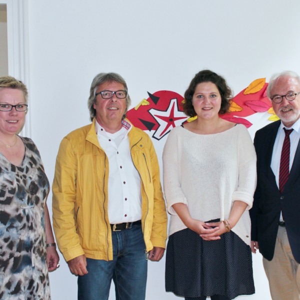 Vier Erwachsene stehen drinnen vor einer weißen Wand mit einem farbenfrohen roten und gelben abstrakten Kunstwerk hinter ihnen. Sie posieren für ein Gruppenfoto und scheinen dabei zu lächeln.