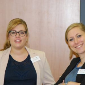 Zwei Frauen in Geschäftskleidung und mit Namensschildern stehen in einem Haus und lächeln in die Kamera. Die eine trägt eine Brille und einen hellen Blazer, die andere einen dunklen Blazer und beugt sich leicht vor.