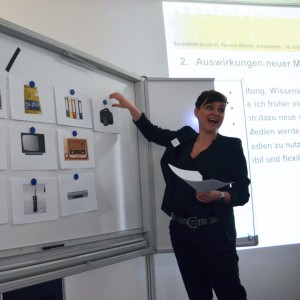Eine Frau steht neben einem Whiteboard, auf dem Bilder verschiedener Geräte, darunter Bücher, Radios, Telefone und Fernseher, zu sehen sind. Sie lächelt und zeigt auf ein Bild, während sie Papiere in der Hand hält, wahrscheinlich hält sie eine Präsentation.