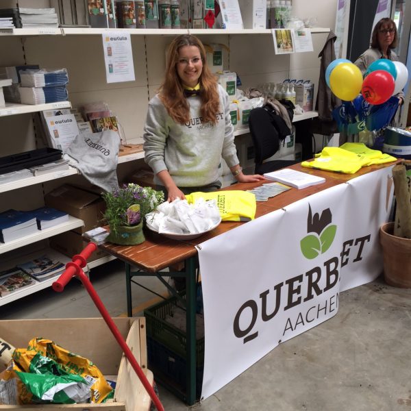 Eine lächelnde Frau steht hinter einem Tisch mit einem Querbeet Aachen-Banner in einem Geschäft. Auf dem Tisch liegen gelbe Hemden, Papiere und Pflanzen. Im Hintergrund befinden sich Regale mit Produkten und eine weitere Person mit Luftballons.