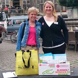 Zwei Frauen stehen lächelnd an einem Informationsstand im Freien mit Flugblättern und einer Tüte mit der Aufschrift "Null Alkohol Voll Power". Im Hintergrund sind ein historisches Gebäude und ein Springbrunnen zu sehen.