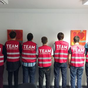 Sieben Personen stehen in einer Reihe vor einer Wand und tragen passende rosa Westen mit der Aufschrift TEAM GLÜCKSSPIELSCHUTZ auf dem Rücken. Über ihnen hängen zwei abstrakte Gemälde an der weißen Wand.