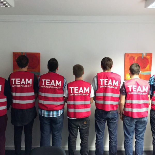 Sieben Personen stehen in einer Reihe vor einer Wand und tragen passende rosa Westen mit der Aufschrift TEAM GLÜCKSSPIELSCHUTZ auf dem Rücken. Über ihnen hängen zwei abstrakte Gemälde an der weißen Wand.