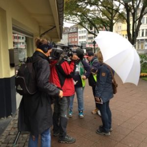 Ein Filmteam mit Kameras nimmt eine kleine Gruppe von Menschen auf, die sich auf einem städtischen Bürgersteig unterhalten; eine Person hält einen großen weißen Regenschirm. Im Hintergrund sind Bäume und Gebäude zu sehen.