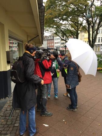 Ein Filmteam mit Kameras nimmt eine kleine Gruppe von Menschen auf, die sich auf einem städtischen Bürgersteig unterhalten; eine Person hält einen großen weißen Regenschirm. Im Hintergrund sind Bäume und Gebäude zu sehen.