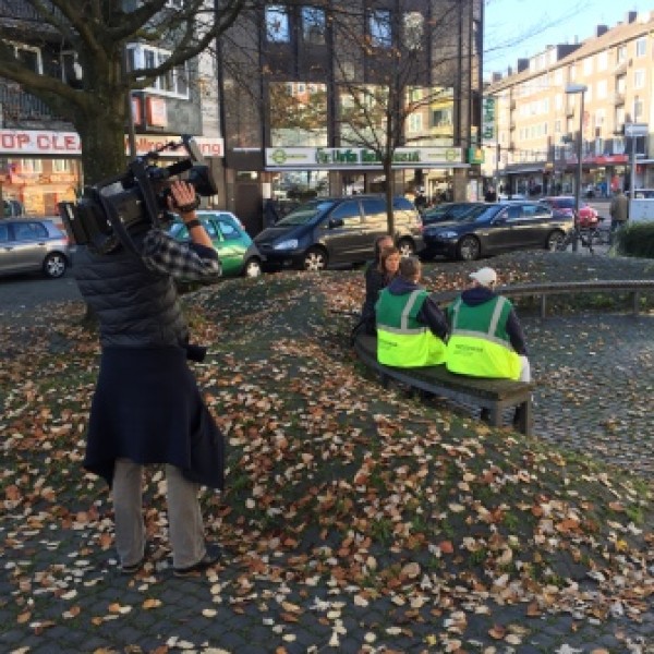 Eine Person bedient eine Videokamera und filmt zwei Personen in grünen Westen, die auf einer Bank in einem begrünten städtischen Gebiet umgeben von parkenden Autos und Gebäuden sitzen. Gefallene Herbstblätter bedecken den Boden.