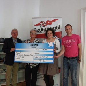 Vier Personen stehen in einem Raum und halten einen großen Präsentationsscheck. Zwei Frauen in der Mitte lächeln, während zwei Männer an den Seiten stehen. Im Hintergrund ist ein Feuervogel-Banner zu sehen.