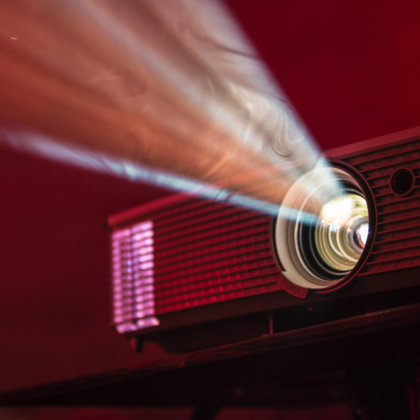 Nahaufnahme eines Projektors in einem dunklen Raum, der einen hellen Lichtstrahl nach vorne wirft, der die Luft vor einem roten Hintergrund in einem dramatischen Licht erhellt.