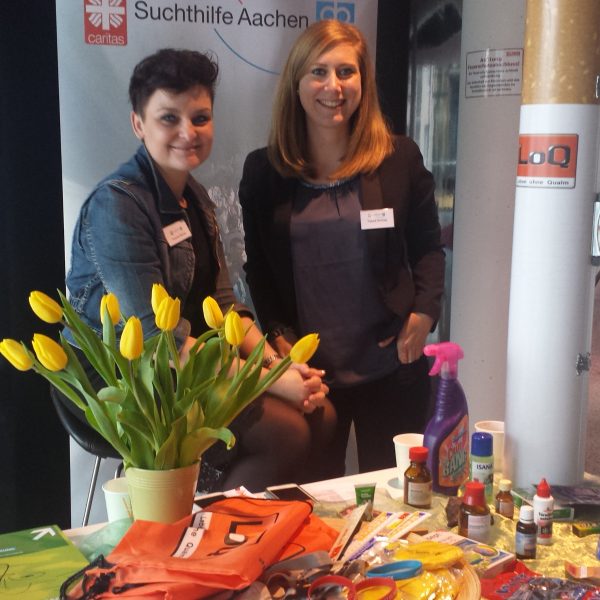 Zwei Frauen stehen hinter einem Schautisch mit Informationsmaterial, Reinigungsmitteln, Snacks und gelben Tulpen. Auf einem Transparent hinter ihnen steht "Suchthilfe Aachen". Sie lächeln in einem überdachten Veranstaltungsraum.