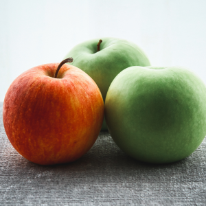Drei Äpfel auf einer grauen, strukturierten Oberfläche; ein roter Apfel links und zwei grüne Äpfel dahinter, vor einem sanft beleuchteten, blassen Hintergrund.