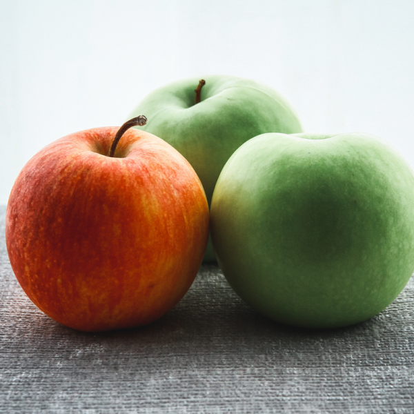 Drei Äpfel auf einer grauen, strukturierten Oberfläche; ein roter Apfel links und zwei grüne Äpfel dahinter, vor einem sanft beleuchteten, blassen Hintergrund.