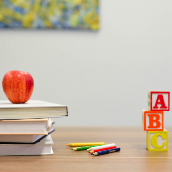 Ein roter Apfel liegt auf einem Bücherstapel neben Buntstiften und Holzklötzen mit den Buchstaben A, B und C auf einem Holztisch. An der hellen Wand im Hintergrund hängt ein verschwommenes Gemälde.
