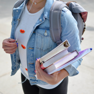 Eine Studentin mit Jeansjacke und Rucksack hält mehrere Bücher und Hefte in einem Arm, während sie auf einem Bürgersteig geht. Ihr Gesicht ist nicht zu sehen.