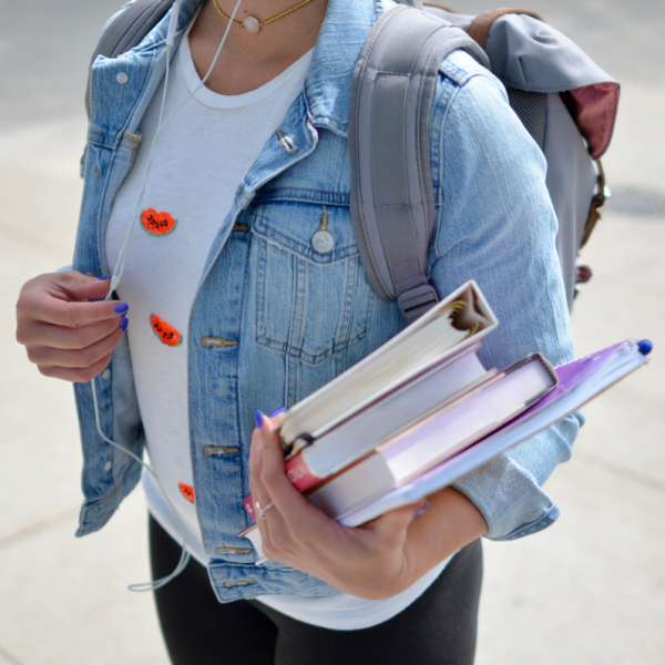 Eine Studentin mit Jeansjacke und Rucksack hält mehrere Bücher und Hefte in einem Arm, während sie auf einem Bürgersteig geht. Ihr Gesicht ist nicht zu sehen.