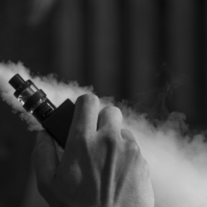 Ein Schwarz-Weiß-Foto in Nahaufnahme von einer Person, die ein Vape-Gerät hält und eine dicke Dampfwolke ausstößt. Nur das untere Gesicht, die Hand und der Dampf sind sichtbar.