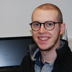 Eine lächelnde Person mit kurzen Haaren, Brille und einem dunklen Pullover mit Reißverschluss sitzt vor einem aufgeklappten Laptop auf einem Schreibtisch.