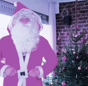 Eine als Weihnachtsmann verkleidete Person in einem rosafarbenen Anzug steht in einem Haus neben einem geschmückten Weihnachtsbaum mit rosafarbenen Ornamenten; im Hintergrund sind eine Backsteinmauer und ein Fenster zu sehen.