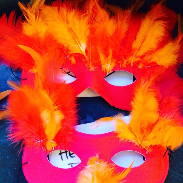 Zwei rote Masken, die mit leuchtend orangefarbenen und gelben Federn verziert sind, liegen übereinander auf einer dunklen Fläche. Die Federn sind voll und flauschig und geben ein feuriges, festliches Aussehen.