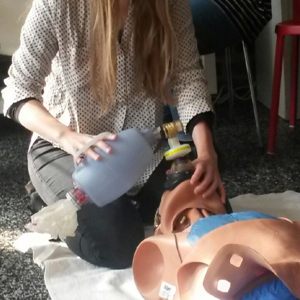 Eine Frau kniet auf dem Boden und übt die Wiederbelebung mit einer Beatmungsbeutelmaske an einer medizinischen Übungspuppe, während eine andere Person im Hintergrund sitzt.