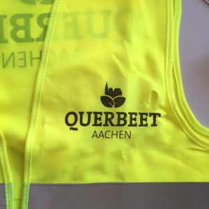 Eine leuchtend gelbe Sicherheitsweste mit einem grauen, reflektierenden Streifen und dem Text "QUERBEET AACHEN" in schwarzer Schrift sowie einem schwarzen Pflanzenlogo oberhalb des Textes.