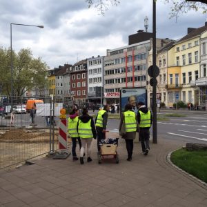 Eine Gruppe von Personen mit gelben Sicherheitswesten geht auf einem städtischen Gehweg neben einer Baustelle entlang und zieht einen kleinen roten Wagen. Bunte Gebäude säumen die Straße im Hintergrund.