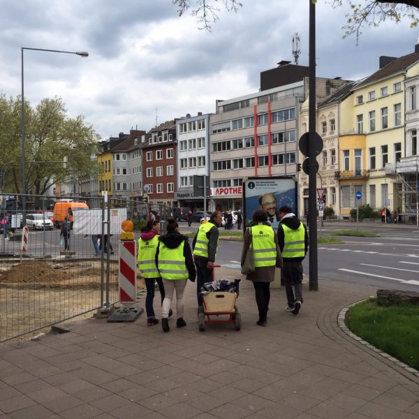 Eine Gruppe von Personen mit gelben Sicherheitswesten geht auf einem städtischen Gehweg neben einer Baustelle entlang und zieht einen kleinen roten Wagen. Bunte Gebäude säumen die Straße im Hintergrund.