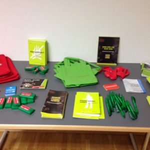 Auf einem Tisch liegen grüne Tragetaschen, grüne Stifte, rote und grüne rechteckige Artikel, Broschüren und rote Kordelzugtaschen, alles fein säuberlich angeordnet für eine Veranstaltung oder Präsentation.