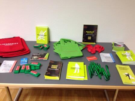 Auf einem Tisch liegen grüne Tragetaschen, grüne Stifte, rote und grüne rechteckige Artikel, Broschüren und rote Kordelzugtaschen, alles fein säuberlich angeordnet für eine Veranstaltung oder Präsentation.