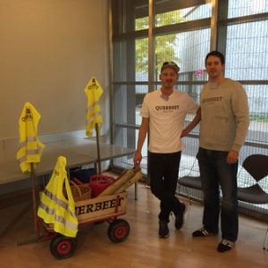 Zwei Personen stehen drinnen neben einem kleinen Holzwagen mit der Aufschrift QUERBEET, der mit Werkzeugen gefüllt und mit gelben Sicherheitswesten bekleidet ist. Beide tragen Hemden mit dem Aufdruck QUERBEET.