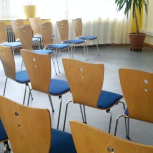 Ein kleiner, leerer Besprechungsraum mit Reihen von Holzstühlen mit blauen Sitzen, einem Schreibtisch mit einem Computer und einem Flipchart auf der linken Seite, großen Fenstern und Topfpflanzen an den Wänden.
