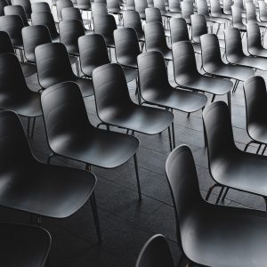 In einem großen Innenraum sind Reihen leerer, moderner schwarzer Stühle ordentlich angeordnet, wodurch ein sich wiederholendes und symmetrisches Muster entsteht. Die Stühle sind aus Kunststoff und haben Metallbeine, und der Boden ist in einer dunklen Farbe gefliest.