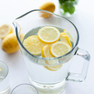 Ein mit Wasser und Zitronenscheiben gefüllter Glaskrug steht auf einer weißen Fläche, daneben zwei leere Gläser, ganze Zitronen und eine kleine Vase mit grünen Minzblättern im Hintergrund.