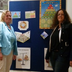 Zwei Frauen stehen lächelnd vor einer blauen Schautafel, die mit Lehrpostern, Tabellen und Diagrammen über Ernährung und gesunde Lebensweise geschmückt ist. Auf der Tafel sind Bilder von Lebensmitteln und Waagen mit deutschem Text zu sehen.