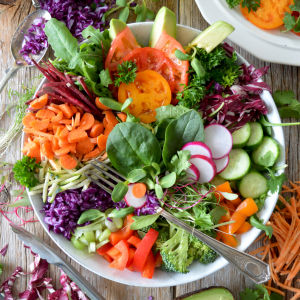Eine bunte Salatschüssel gefüllt mit Spinat, Tomaten, Avocado, Karotten, Radieschen, Gurken, Rotkohl und Paprika. Um die Schüssel herum stehen Salatzutaten, Utensilien, Olivenöl und Avocado auf einem rustikalen Holztisch.